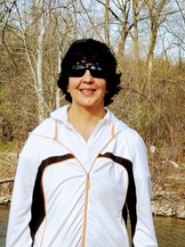 Bharati is a South-Asian female with short black hair and light brown skin colour. She is wearing a white shirt, a lightweight summer outdoor jacket with orange full zip and a white hoodie, and dark square sunglasses with black frames. She is standing in a wooded area with trees and water. 