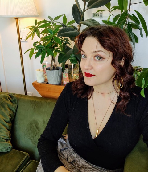 Megan, a white disabled femme with brown hair and green eyes slyly stares at the camera from the side. She is wearing red lipstick, black, and topped off with big gold jewellery and sitting comfortably on a green velvet couch, surrounded by plants. 