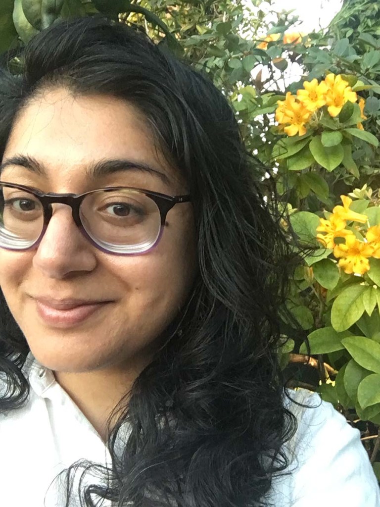 A close up of a South Asian femme wearing a white collared shirt and large rounded glasses. Sarah is smiling and slightly out of frame, standing in front of a large shrub with clusters of yellow flowers.